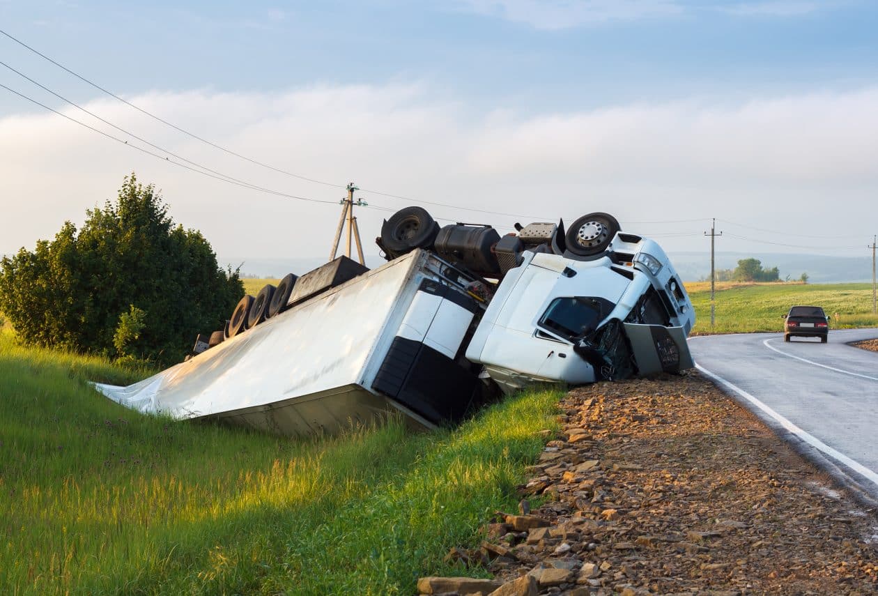 Camión volcado en el arcén de una carretera rural, con la cabina y el remolque completamente fuera de la vía. El accidente ocurrió en una curva, y un coche se aleja por la carretera. El paisaje circundante es verde con colinas y postes eléctricos visibles.